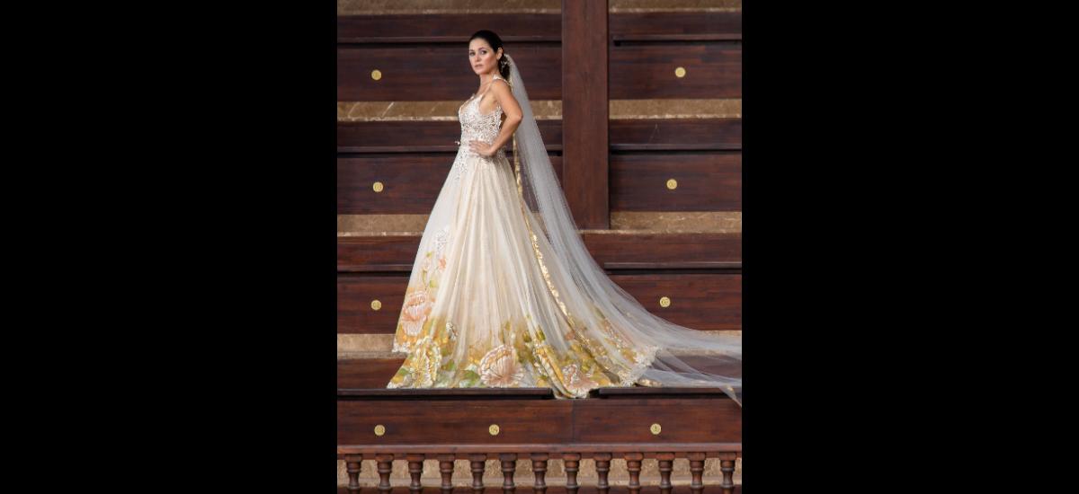 THALÍA SEGRERA: Vestido de novia ajustable a las diferentes etapas de la gran fecha. La primera etapa es un vestido corte princesa con falda y cola desprendibles, pintadas a mano con oleo interpretado por artistas plásticos sobre chiffón metalizado dorado, bordado con apliques en guipur, perlas y cristales. La parte superior se representa en un escote en V con tiras en guipur beige bordado con cristales y perlas. El velo es pintado a mano con lamilla de oro. Para la segunda etapa del vestido se quita el la cola y el pollerín, dejando ver en la trasparencia de la falda y un enterizo de pantalón en el mismo guipur, para un look más relajado. La última etapa se desprende de la falda y queda en un enterizo de guipur bordado en perlas y cristales, con abertura en los costados, y la cola del vestido pasa a ser una capa en la espalda; y como accesorio, una corona de flores con luces.